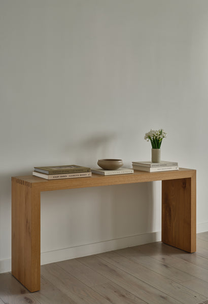 The Malibu Console Table in White Oak stands against a plain white wall, displaying box joint details. Stacked books, a beige bowl, and a small vase with white flowers complete its clean, modern style on the light wood floor.