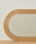 A close-up of the Oak Oval Mirror, showcasing its modern solid wood frame with rounded rectangular edges in light white oak tones, set against a plain off-white wall and a wooden surface.