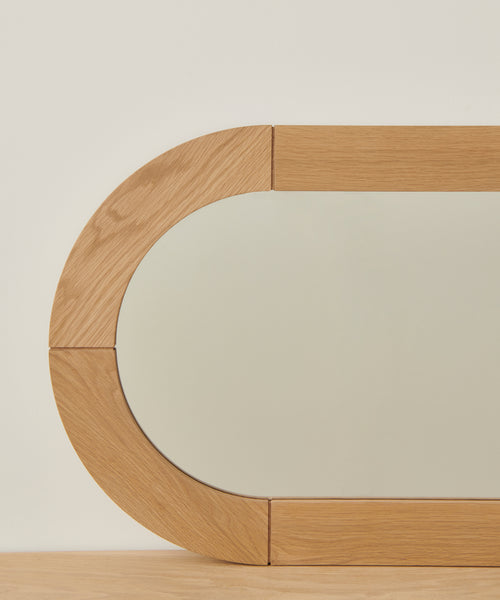 A close-up of the Oak Oval Mirror, showcasing its modern solid wood frame with rounded rectangular edges in light white oak tones, set against a plain off-white wall and a wooden surface.