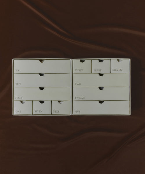 The Oak Essentials x Jenni Kayne Advent Calendar features two open boxes with small drawers numbered one to twelve, arranged randomly on a brown, silky fabric—ideal for advent surprises or thoughtful self-care gifts.