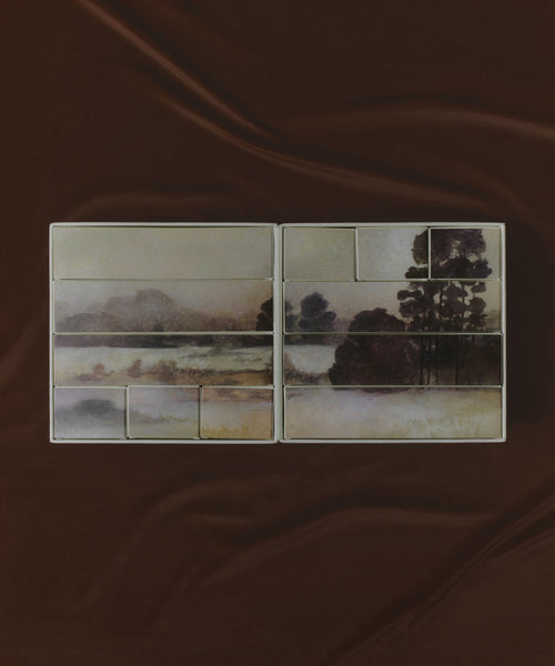 The Oak Essentials x Jenni Kayne Advent Calendar features rectangular palettes with powder pans adorned by misty landscape art of trees and water, elegantly displayed on dark brown fabric—an ideal self-care gift.