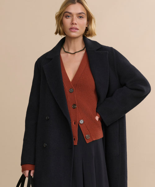 A woman with short blonde hair wears the oversized Olivia Coat over a rust-colored buttoned cardigan and black skirt, holding a black bag, standing against a plain beige background.