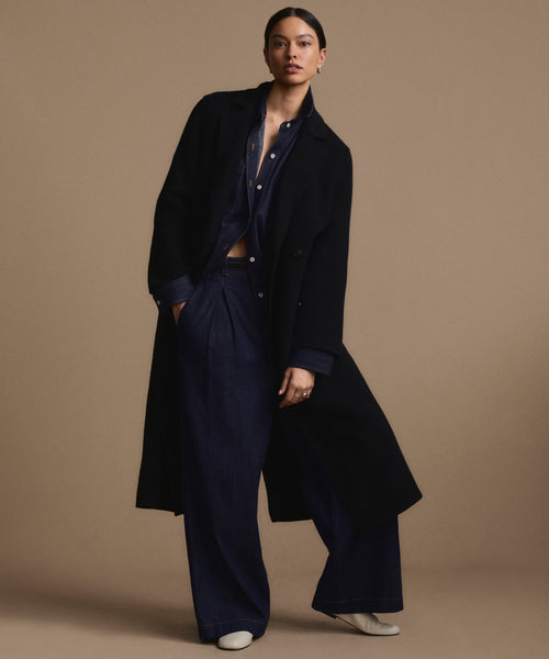 A person poses against a beige background in an oversized wool Olivia Coat, layered over a navy shirt and wide-leg pants with white shoes. Their left hand rests in their pocket, and they maintain a neutral expression.