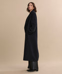 A woman with shoulder-length brown hair stands in profile against a beige backdrop, wearing the oversized Olivia Coat, wide-leg black pants, and black shoes, her hands tucked into her pockets—a timeless women's outerwear look.