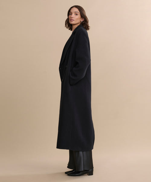 A woman with shoulder-length brown hair stands in profile against a beige backdrop, wearing the oversized Olivia Coat, wide-leg black pants, and black shoes, her hands tucked into her pockets—a timeless women's outerwear look.