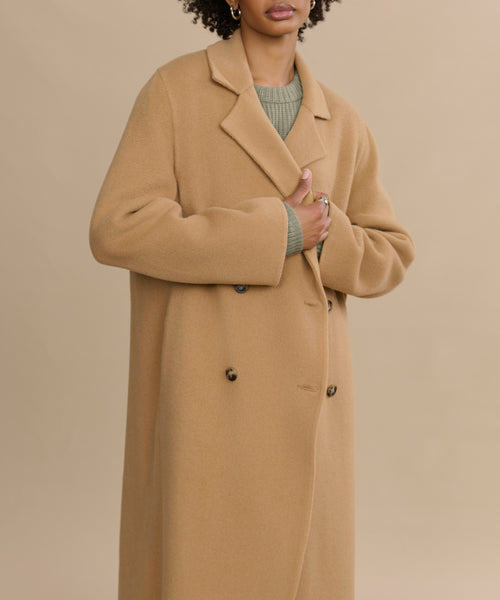 Someone wears the Olivia Coat—an essential oversized camel wool outerwear piece—over a green sweater, standing against a beige background. Their hand holds the coat closed, with their face partially out of frame.