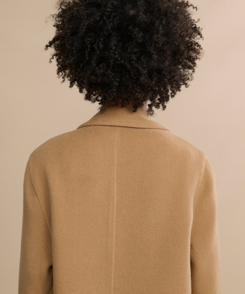 A person with short, curly dark hair is shown from behind, wearing the Olivia Coat—a stylish camel wool outerwear—set against a beige background.