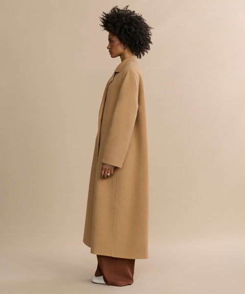 A woman stands in profile against a neutral background, wearing the oversized Olivia Coat—an outerwear essential—in camel wool over brown pants and white shoes.