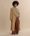 A woman with curly hair poses confidently against a beige background, wearing the oversized Olivia Coat in camel wool over a green sweater, wide brown trousers, and white shoes, with one hand casually placed in her pocket.