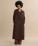 A woman with curly hair stands against a plain beige background, hands in the pockets of her long, chocolate Olivia Coat, layered over brown pants and a brown top.