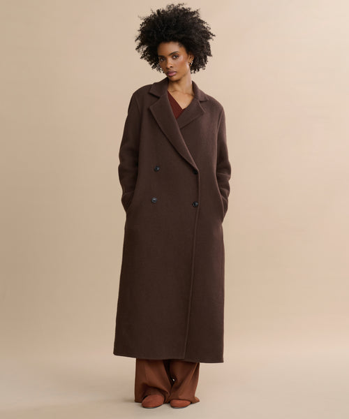 A woman with curly hair stands against a plain beige background, hands in the pockets of her long, chocolate Olivia Coat, layered over brown pants and a brown top.