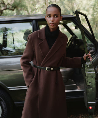 A woman wearing the Olivia Coat, a long, chocolate oversized wool coat with a black belt, stands by an open car door looking to the side. Lush trees and greenery can be seen in the background.