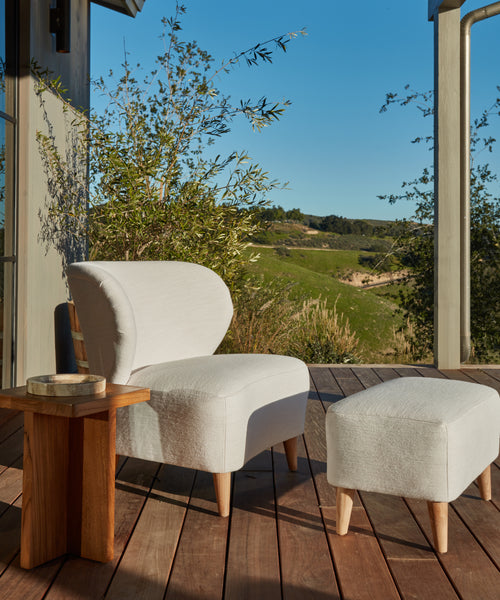 A modern white armchair with a matching Outdoor Brentwood Ottoman sits on a wooden deck beside a small teak table. Green hills and blue sky appear in the background, sunlight casting gentle shadows over the scene.