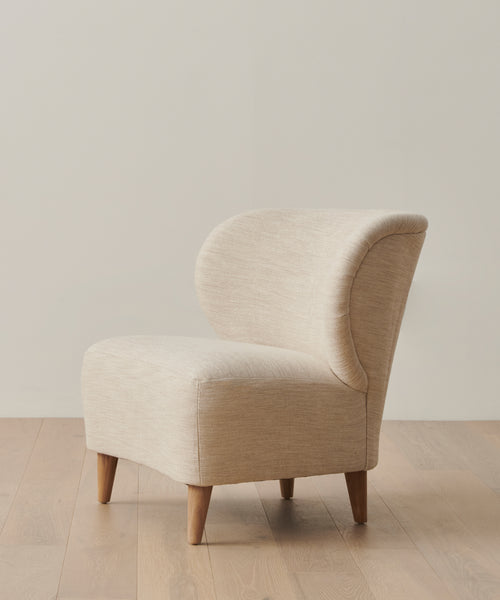The Outdoor Brentwood Chair, featuring light beige performance textured linen upholstery and solid teak legs, sits on a light wood floor against an off-white wall.