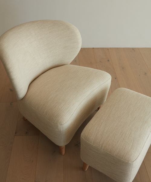 The Outdoor Brentwood Chair, with a modern beige upholstered design, curved backrest, and solid teak legs, sits beside a matching rectangular ottoman on a light wooden floor, both in performance textured linen.