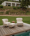Two cream-colored lounge chairs with solid teak legs and an Outdoor Brentwood Ottoman are set on a wooden poolside deck, separated by a small wooden side table. A modern house and grassy hillside appear in the background.