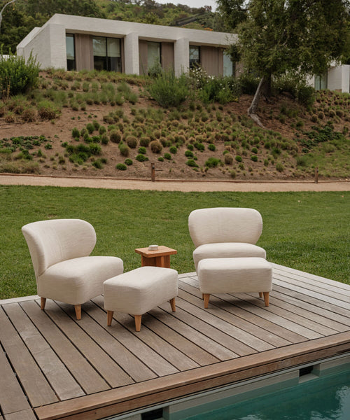 Two cream-colored lounge chairs with solid teak legs and an Outdoor Brentwood Ottoman are set on a wooden poolside deck, separated by a small wooden side table. A modern house and grassy hillside appear in the background.
