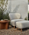 A modern outdoor space showcases the Outdoor Brentwood Chair with solid teak legs and light gray cushions, paired with a matching ottoman on gravel beside a wooden table, surrounded by lush plants and a light-colored wall.