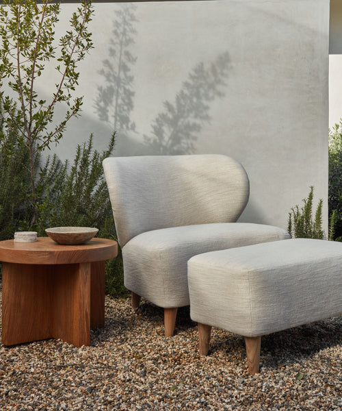 A modern outdoor space includes a light gray cushioned chair with solid teak legs and the Outdoor Brentwood Ottoman on gravel, beside a wooden side table with ceramic bowls, all set amid greenery against a pale wall.