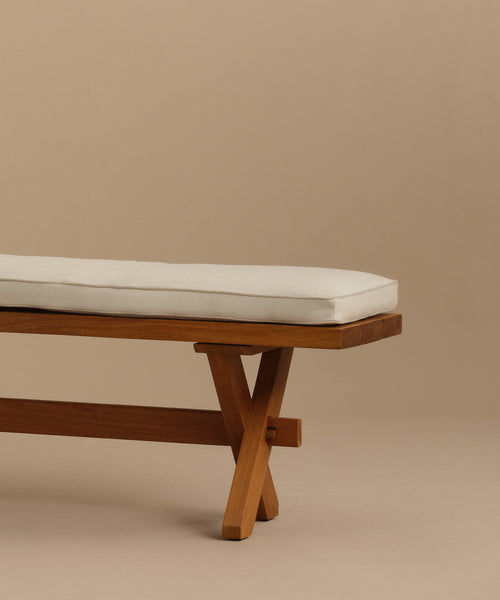 The Outdoor Garden Dining Bench, featuring solid teak and X-shaped legs with a Tempotest fabric cushion, sits partially visible against a plain beige background, emphasizing its minimalist and modern style.