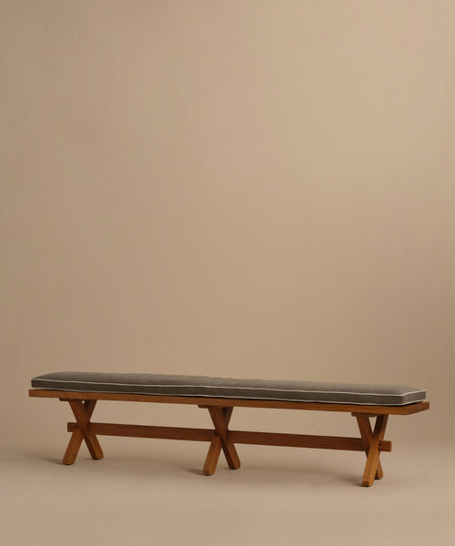 The Outdoor Garden Dining Bench features a long solid teak frame, cushioned seat in dark gray Tempotest fabric, and crossed wooden legs, shown against a plain beige background.