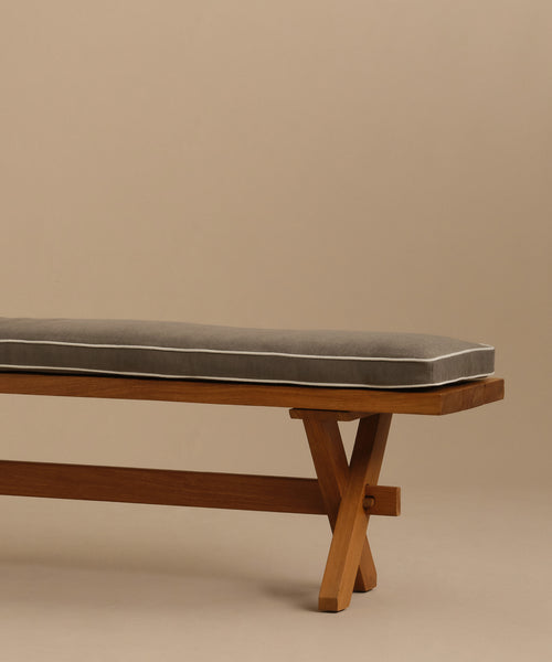 The Outdoor Garden Dining Bench features crossed solid teak legs and a gray Tempotest fabric cushion, shown against a plain beige background.