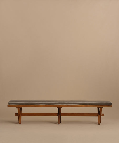 The Outdoor Garden Dining Bench features a minimalist solid teak design topped with a slim, dark Tempotest fabric cushion, shown against a plain beige background.