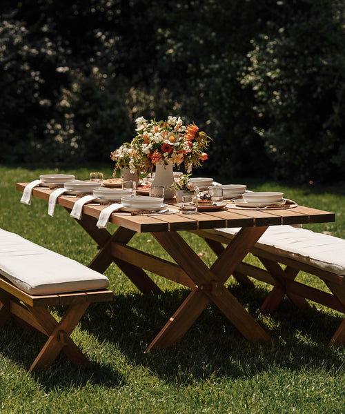 The Outdoor Garden Dining Table is arranged with white dishes, glasses, and napkins. A floral centerpiece adds charm as the table sits on green grass with trees in the background. Matching benches complete the garden dining setup.