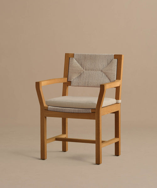 The Outdoor Montecito Dining Arm Chair features solid teak, a light beige cushioned seat, and a textured patterned fabric backrest, displayed against a plain beige background.