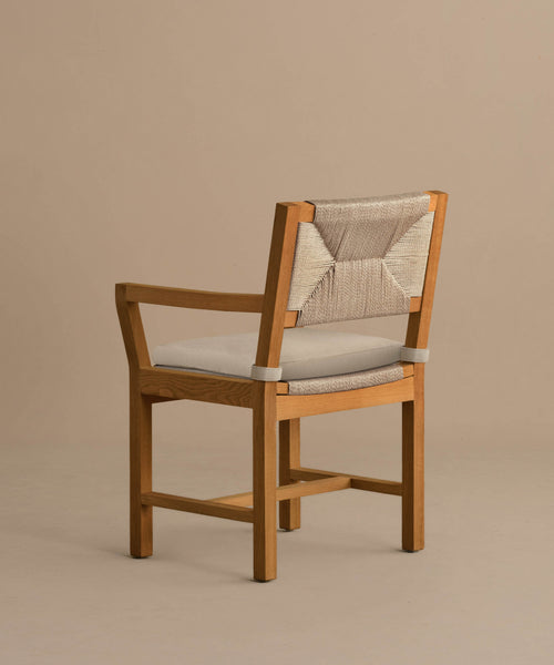 The Outdoor Montecito Dining Arm Chair features a solid teak frame, a beige cushioned seat, and a woven beige backrest in a minimalist, modern design, shown against a plain light brown background.