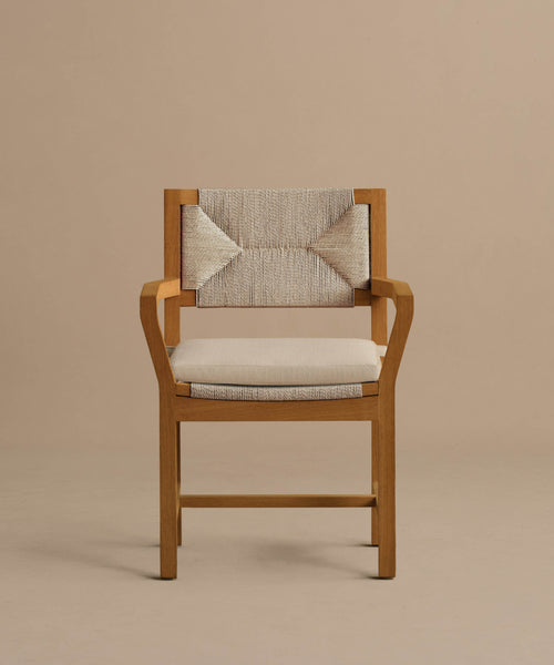 The Outdoor Montecito Dining Arm Chair features a solid teak frame, angled armrests, a beige cushioned seat, and a textured woven backrest with a decorative bow accent, shown against a plain taupe background.