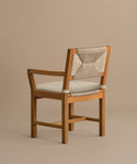 The Outdoor Montecito Dining Arm Chair features solid teak, a light beige Tempotest fabric cushion, and a woven backrest. Its simple, modern design highlights the natural wood grain and clean lines against a taupe background.