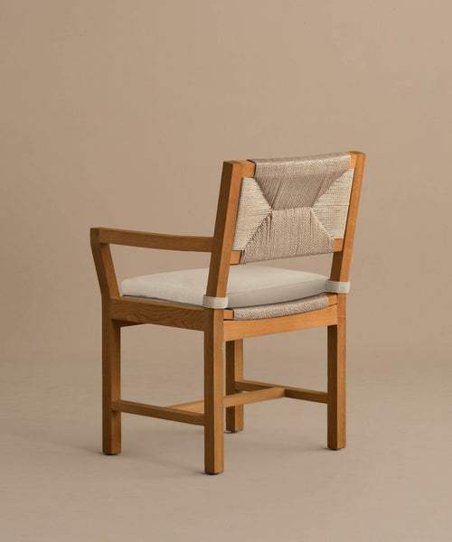 The Outdoor Montecito Dining Arm Chair features solid teak, a light beige Tempotest fabric cushion, and a woven backrest. Its simple, modern design highlights the natural wood grain and clean lines against a taupe background.