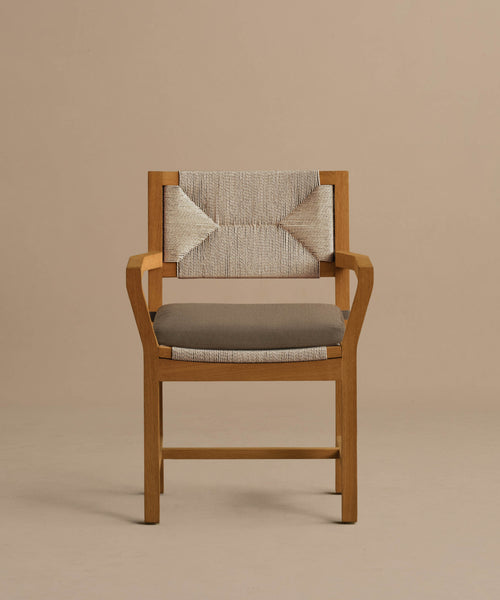 The Outdoor Montecito Dining Arm Chair features a solid teak frame, a woven ECOLENE beige backrest, and a gray cushioned seat. Its simple angular armrests and legs offer a sleek, modern design against a plain beige background.