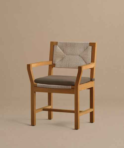 The Outdoor Montecito Dining Arm Chair features a solid teak frame, beige textured fabric on the back, and a dark gray cushioned seat, shown against a plain beige background.