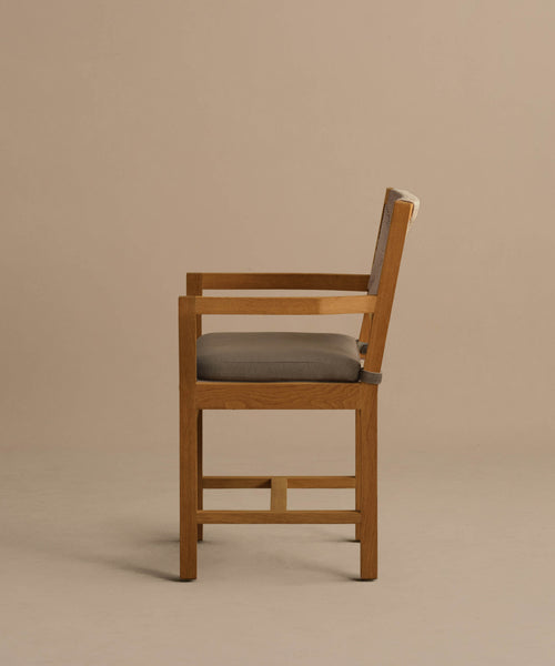 The Outdoor Montecito Dining Arm Chair, crafted from solid teak with a straight back and square frame, features cushioned seat and backrest in light gray fabric, shown in a side view against a plain beige background.