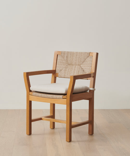 The Outdoor Montecito Dining Arm Chair Cover protects your wooden armchair with a woven backrest and light beige cushion, helping it stay looking new year-round on any wooden floor against an off-white wall.