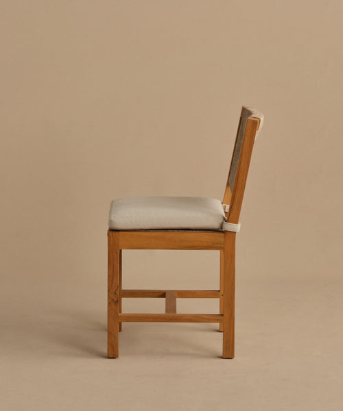 The Outdoor Montecito Dining Chair features a teak frame and light beige cushioned seat and backrest. Shown in profile against a plain, light brown background, its modern design offers style and durability for outdoor dining spaces.