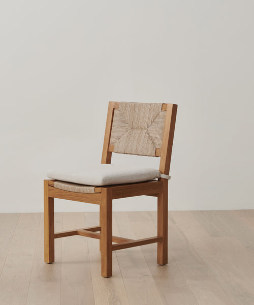 A wooden chair with a woven backrest and light cushion sits on a wood floor by an off-white wall—perfectly protected for lasting beauty with the Outdoor Montecito Dining Chair Cover, which is UV resistant.