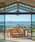 An Outdoor Montecito Oval Dining Table with chairs is set on a patio overlooking rolling green hills and distant mountains, framed by large open glass doors under a modern pergola roof.