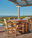 An Outdoor Montecito Oval Dining Table with six chairs sits on a deck under a pergola, facing scenic green hills and distant water beneath a clear blue sky.