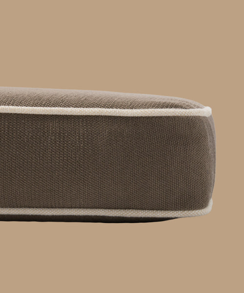 A close-up side view of the Outdoor Vista Chair Cushion, a brown rectangular cushion with white piping made from Italian fabric, set against a plain beige background.
