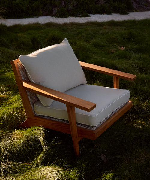 The Outdoor Vista Chair, made of modern teak and light gray Tempotest fabric cushions, is placed on vibrant green grass outdoors with blurred foliage and a dirt path in the background.