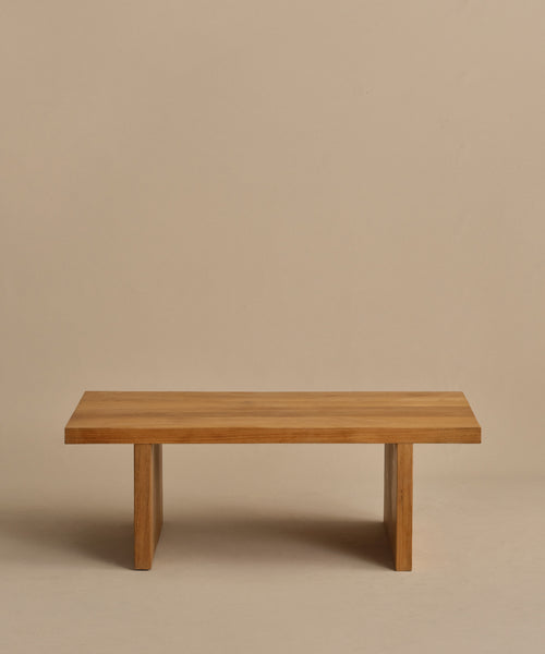 The Outdoor Vista Coffee Table, a minimalist teak piece with a rectangular top and two wide, flat legs, is shown against a plain beige background.