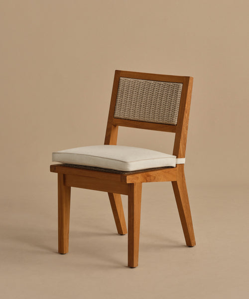 The Outdoor Vista Dining Chair features solid teak construction, angled legs, a woven rattan backrest, and a thick white seat cushion, shown against a plain beige background.