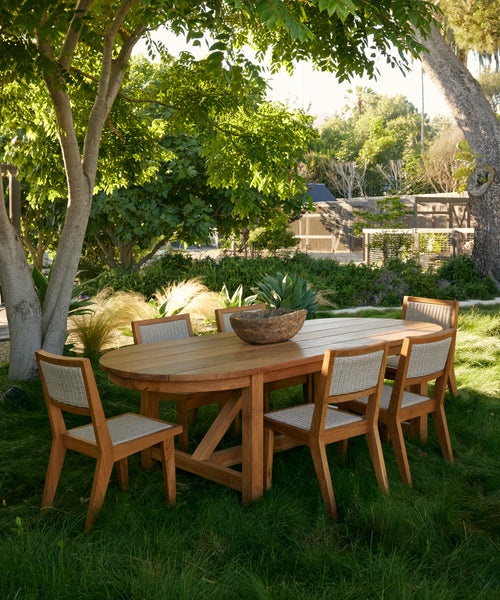 Six Outdoor Vista Dining Chairs and a wooden outdoor dining table are arranged on green grass beneath leafy trees in a lush garden. A large decorative bowl sits on the table, surrounded by plants and sunlight.