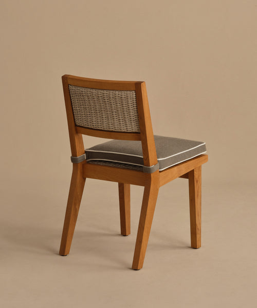 The Outdoor Vista Dining Chair features a solid teak frame, woven ECOLENE backrest, and gray cushioned seat. Shown at an angle on a plain beige background to highlight its back and side details.