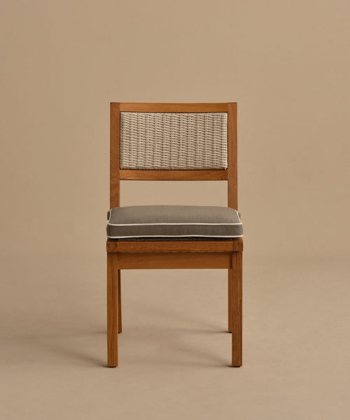 The Outdoor Vista Dining Chair features solid teak construction, a woven rattan backrest, and a gray seat cushion, shown against a plain beige background.