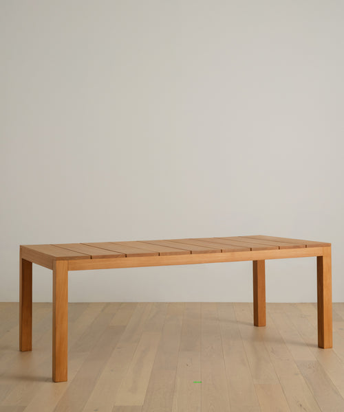 The Outdoor Vista Dining Table, featuring a rectangular solid teak top and four straight legs, sits on a light wood floor against a plain, light-colored wall.