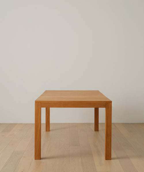 The Outdoor Vista Dining Table, a simple rectangular solid teak design with four legs, stands on a light wood floor against an off-white wall for a minimalistic and uncluttered look.
